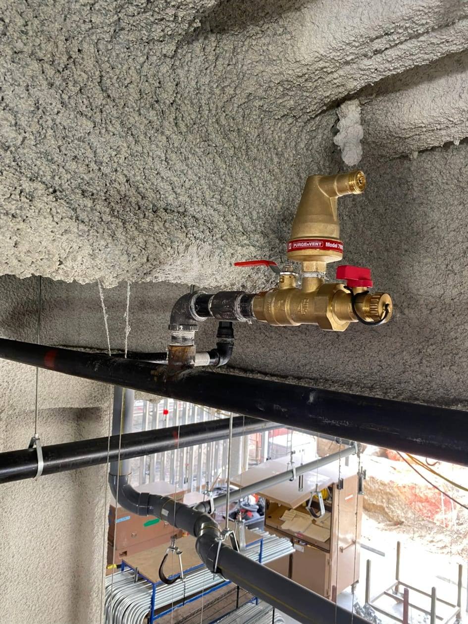 Brass air relief vent and ball valve assembly installed on a fire sprinkler main beneath a sprayed fireproofed deck