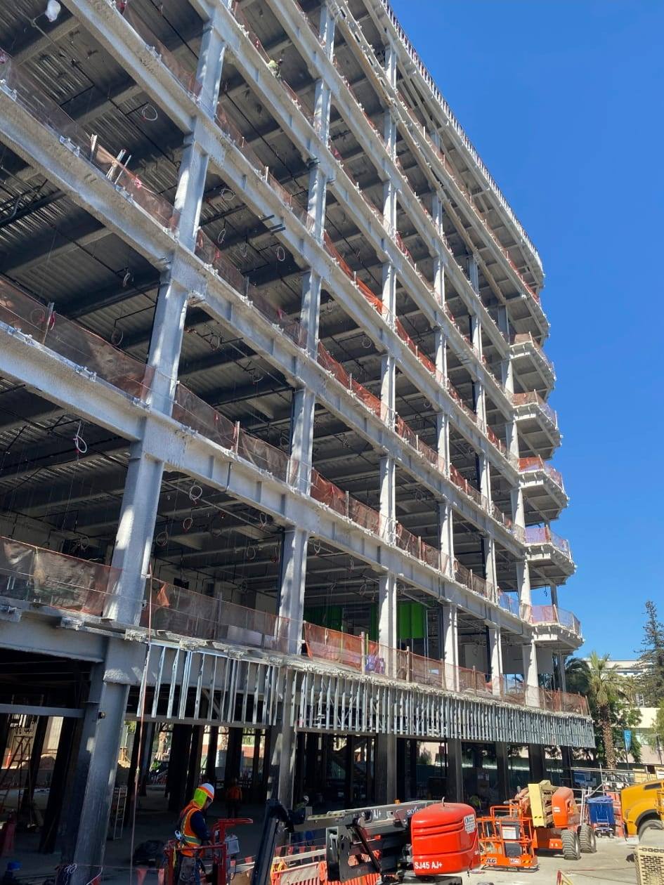Concrete building under construction with fire sprinkler drops visible on every floor, boom lifts and a worker in a high-visibility vest at ground level