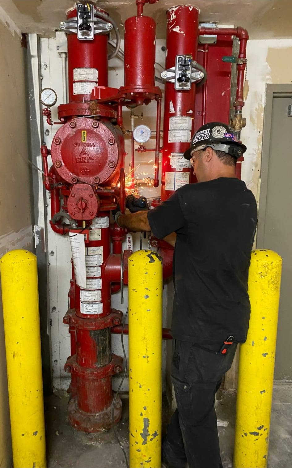 Technician in a hard hat cutting into a red Grinnell dry-pipe riser valve assembly, sparks flying, behind yellow bollards in a mechanical room