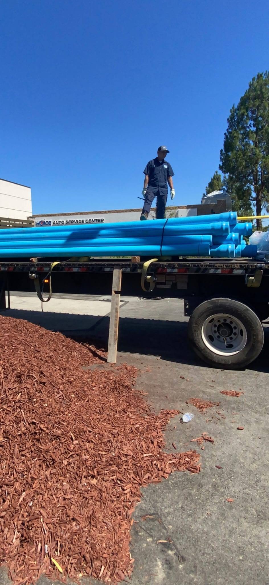 Flatbed truck loaded with lengths of blue underground fire main pipe, a crew member in uniform standing on top of the load under a clear sky