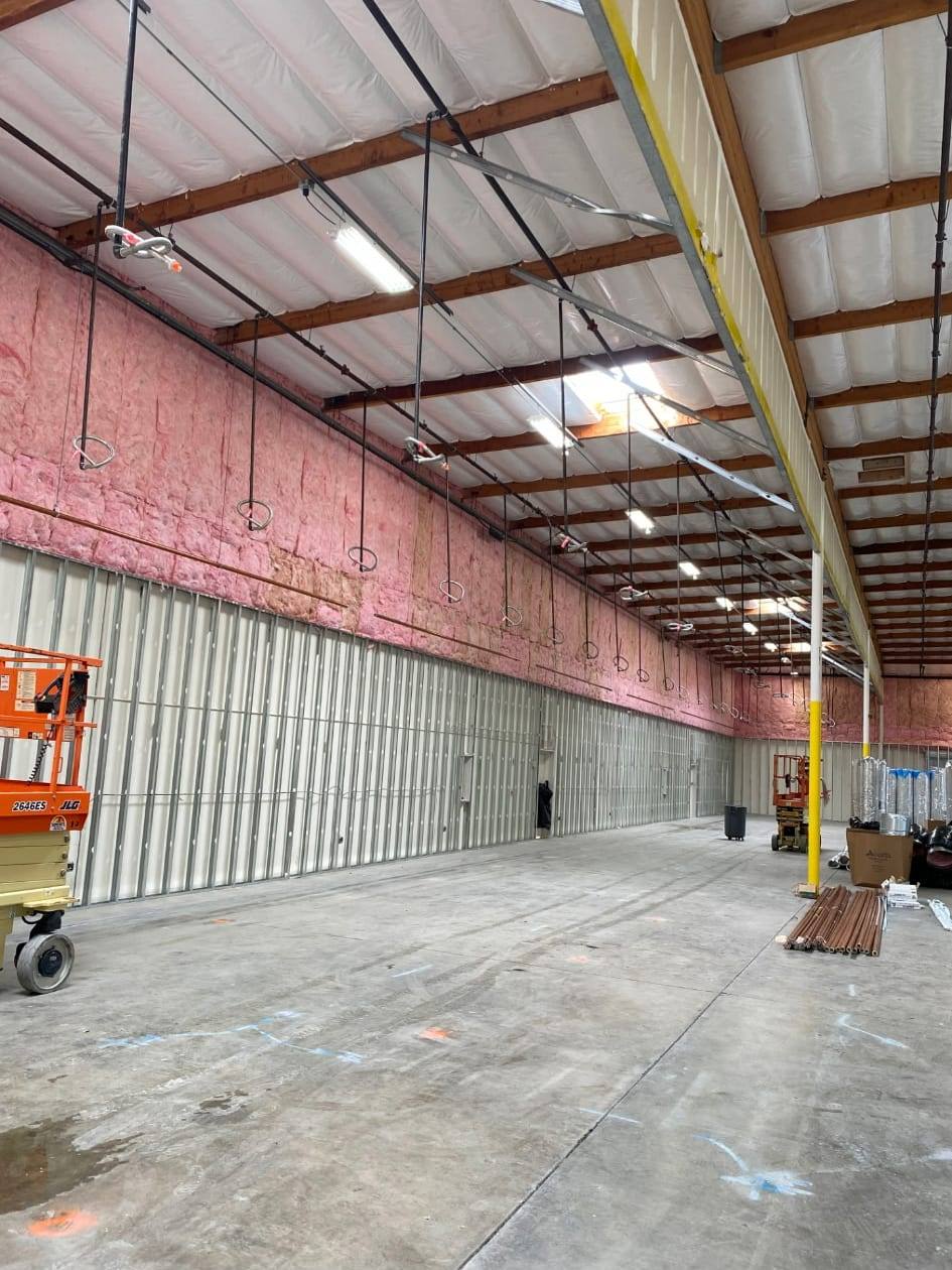Interior of a large insulated warehouse with rows of fire sprinkler drops hanging from the roof structure and a scissor lift parked against the wall